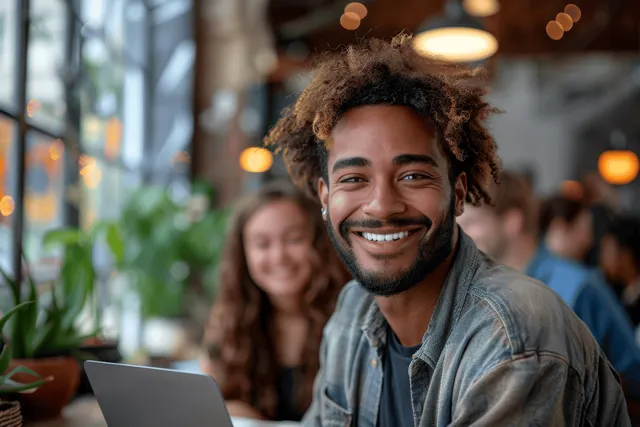 Person working on a laptop at an office desk with blurred coworkers and warm lights behind.