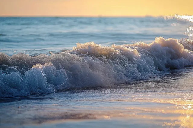 Close-up ocean waves break on sandy shore under golden sunset sparkle and calm horizon