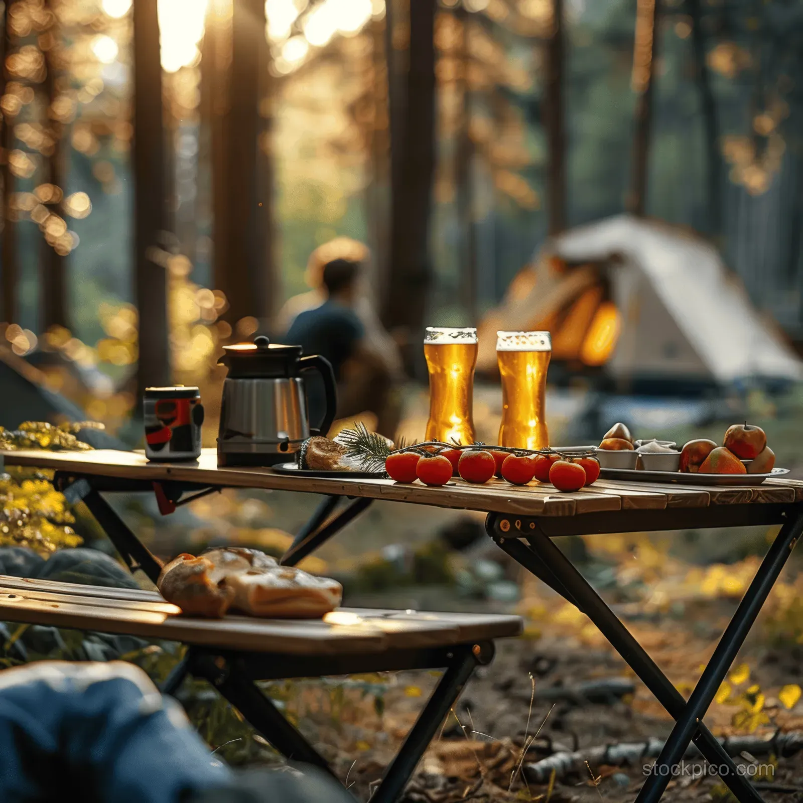 Camping table outdoors with coffee pot, beer glasses, and fruit plate near a forest tent at sunrise.