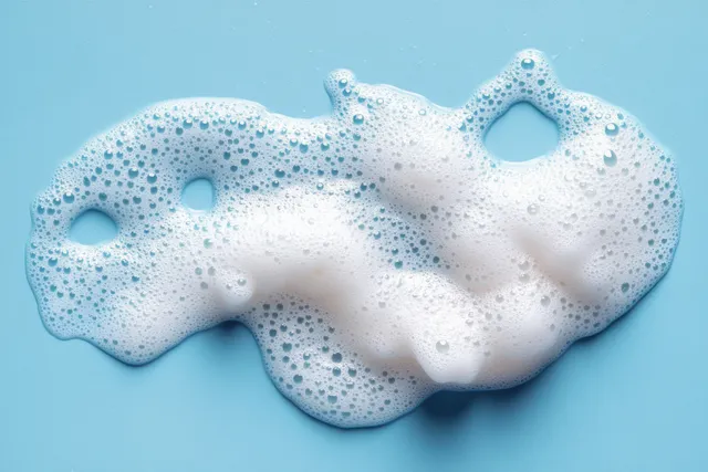 Lathered facial cleanser mousse foam rests on a blue background with many small bubbles visible.