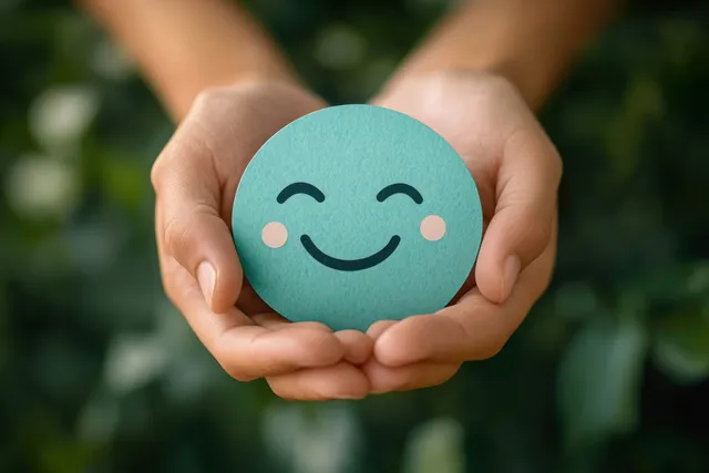 Hands gently cradle a round teal smiling face card against a softly blurred green outdoor background.