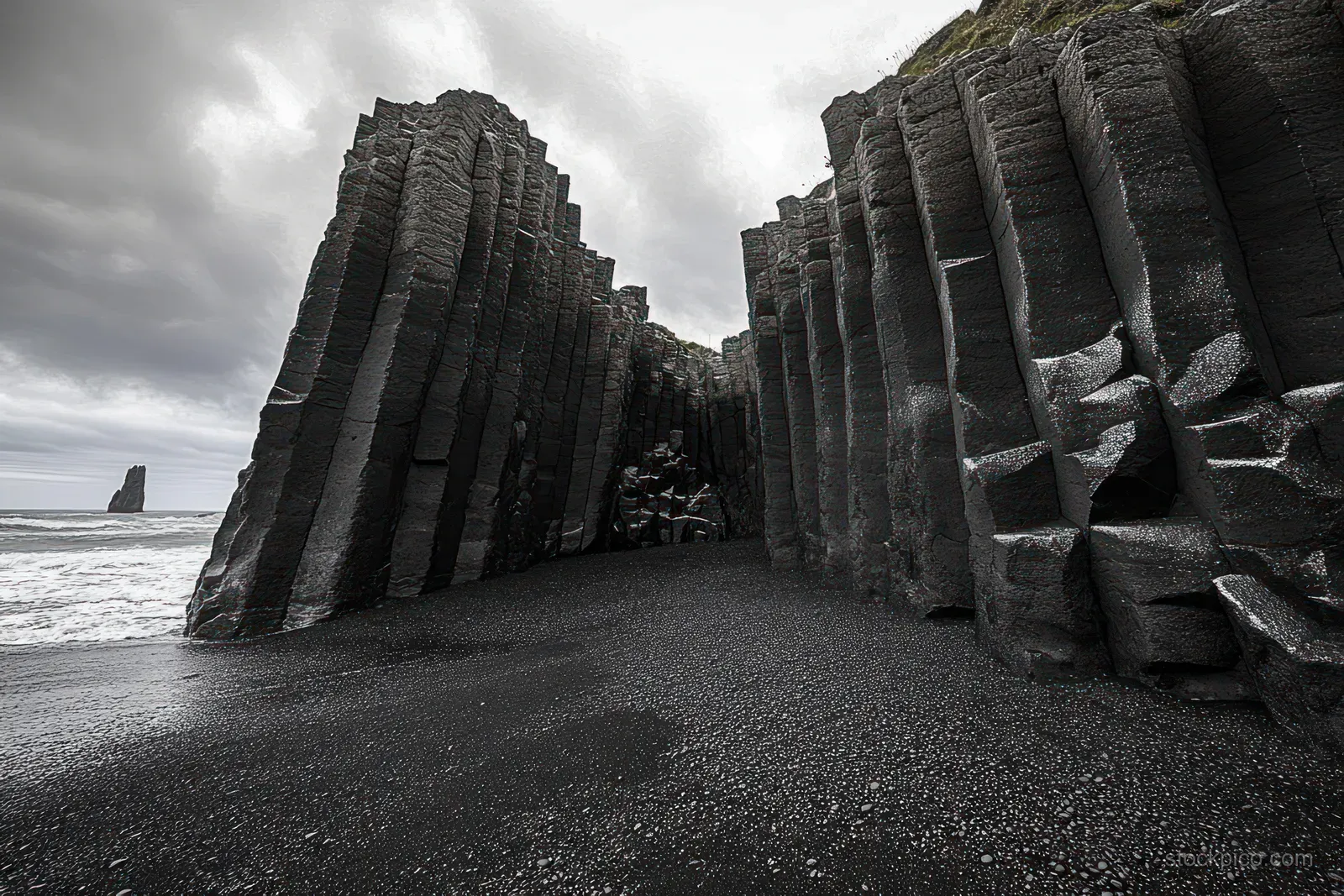 Hexagonal basalt columns stand on a black sand beach with ocean waves and cloudy storm skies overhead.