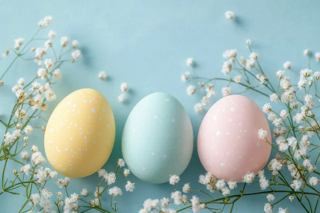 Three pastel speckled eggs in yellow, blue, and pink surrounded by white baby's breath on a soft blue background.