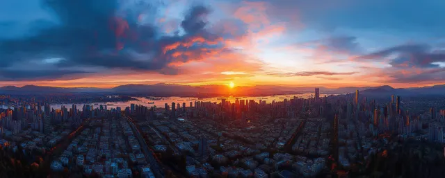Panoramic sunset cityscape over waterfront, dense high-rises, distant mountains, and colorful clouds