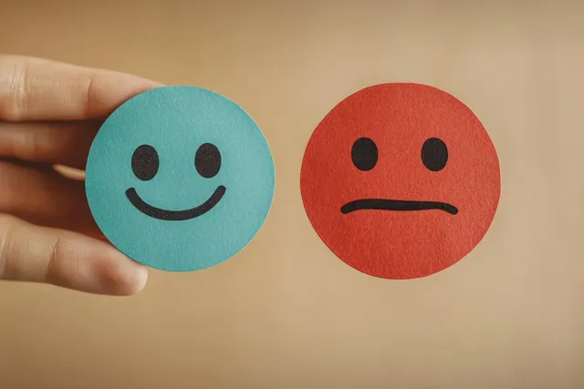 A hand holds a blue smiley paper circle beside a red frowning face circle on a beige background.
