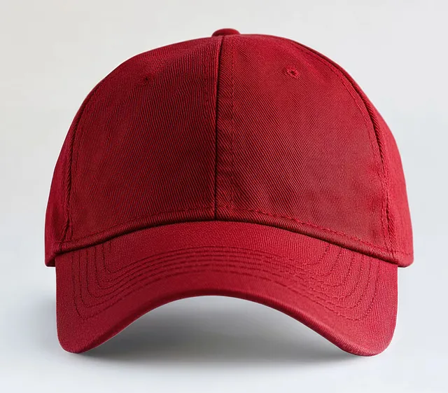 Plain red baseball cap close-up showing panel stitching and curved brim on a light neutral background.