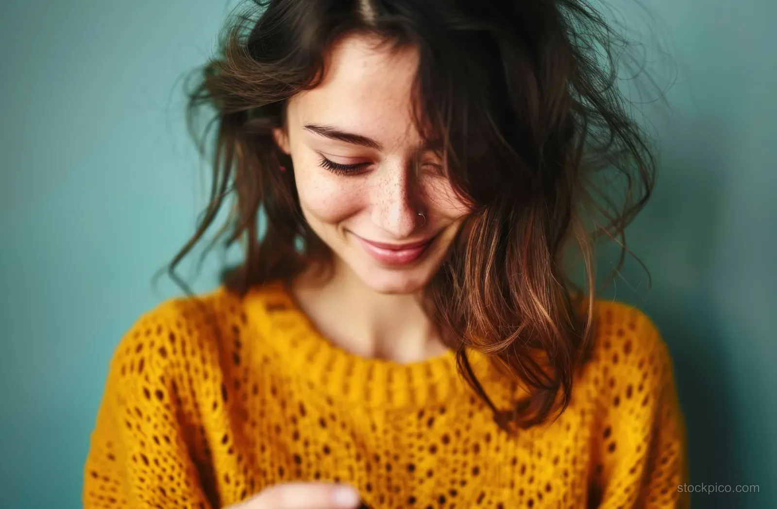 Woman with long wavy hair wearing a mustard knitted sweater, smiling against a teal background.