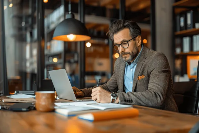An accountant reviews invoices and notes on a laptop at a modern office desk under warm lights.