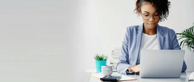 Businesswoman typing on a laptop at her desk next to a calculator and notebook with finance papers.