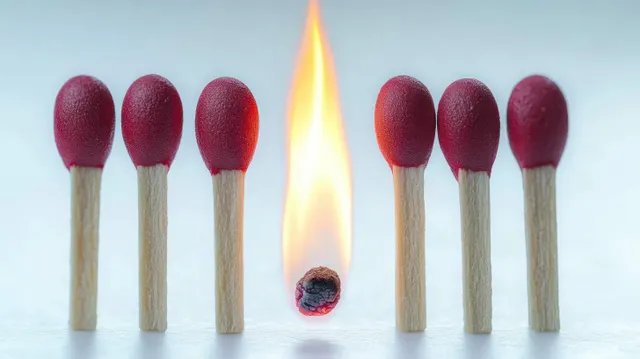 Six red matchsticks surround one burning matchstick with a bright flame on a light background.
