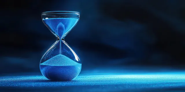 A transparent hourglass pours blue sand onto a blue surface under dramatic dark lighting.