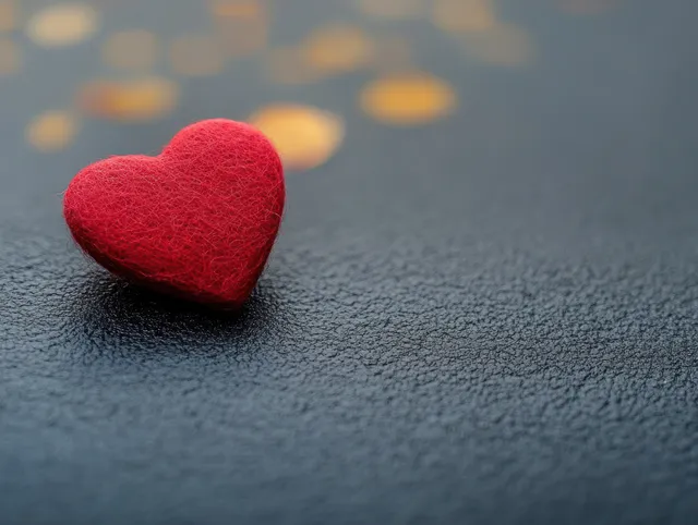Small red felt heart rests on a dark textured surface with blurred golden bokeh in the background.