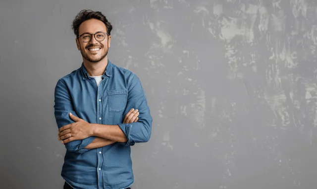 Smiling man in a blue shirt and glasses stands with arms crossed next to grey copy space.