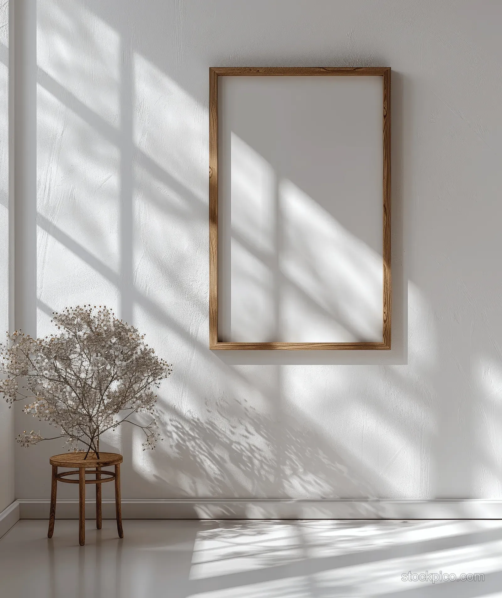 Vertical wooden picture frame mockup mounted on a white wall with window light shadows and a small wooden stool beside it.