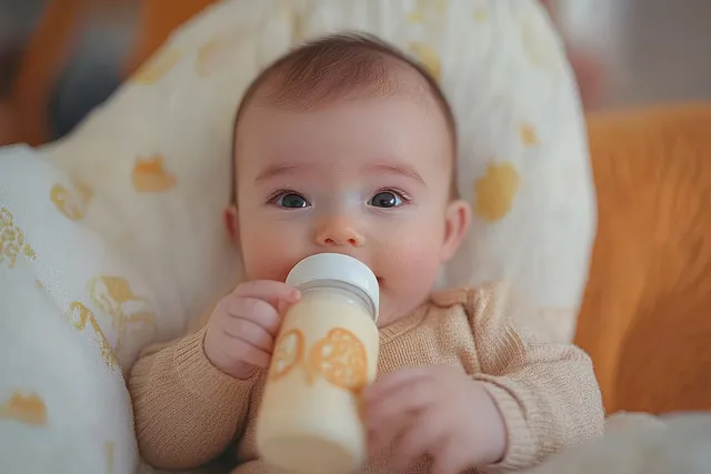 A baby lies on a patterned blanket drinking from a bottle and making eye contact with the camera.