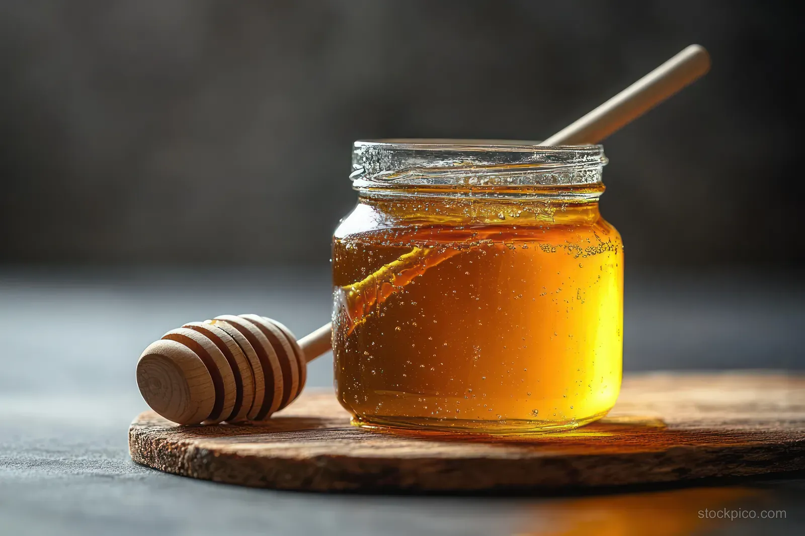 A glass jar of golden honey with a wooden honey dipper resting on a rustic wooden board under warm light.