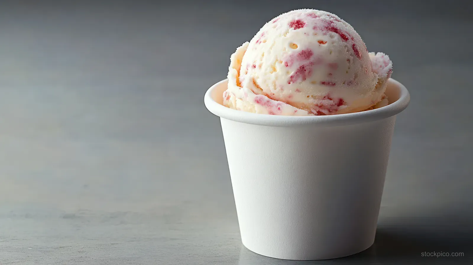 A single scoop of strawberry swirl ice cream in a white paper cup on a smooth gray surface.