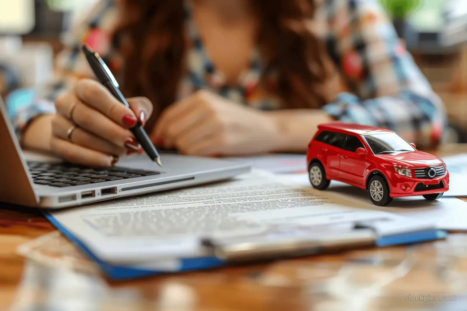 A woman signs a car deal contract at a desk with an open laptop and a red SUV model nearby.