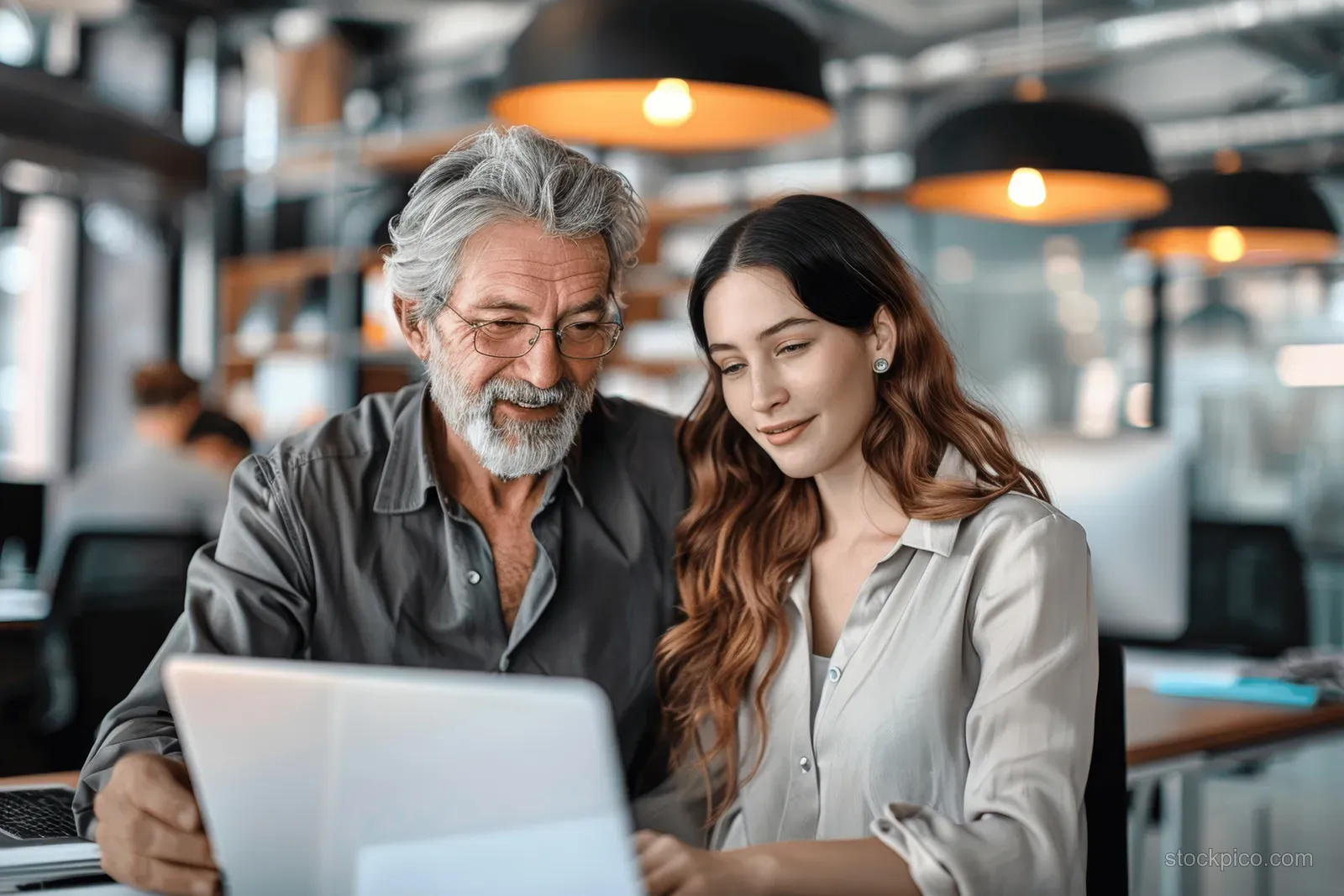 An older man and younger woman discuss results on a laptop in a bright coworking setting with warm lights.