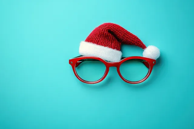 Red-framed glasses wearing a small Santa hat rest on a bright blue background.