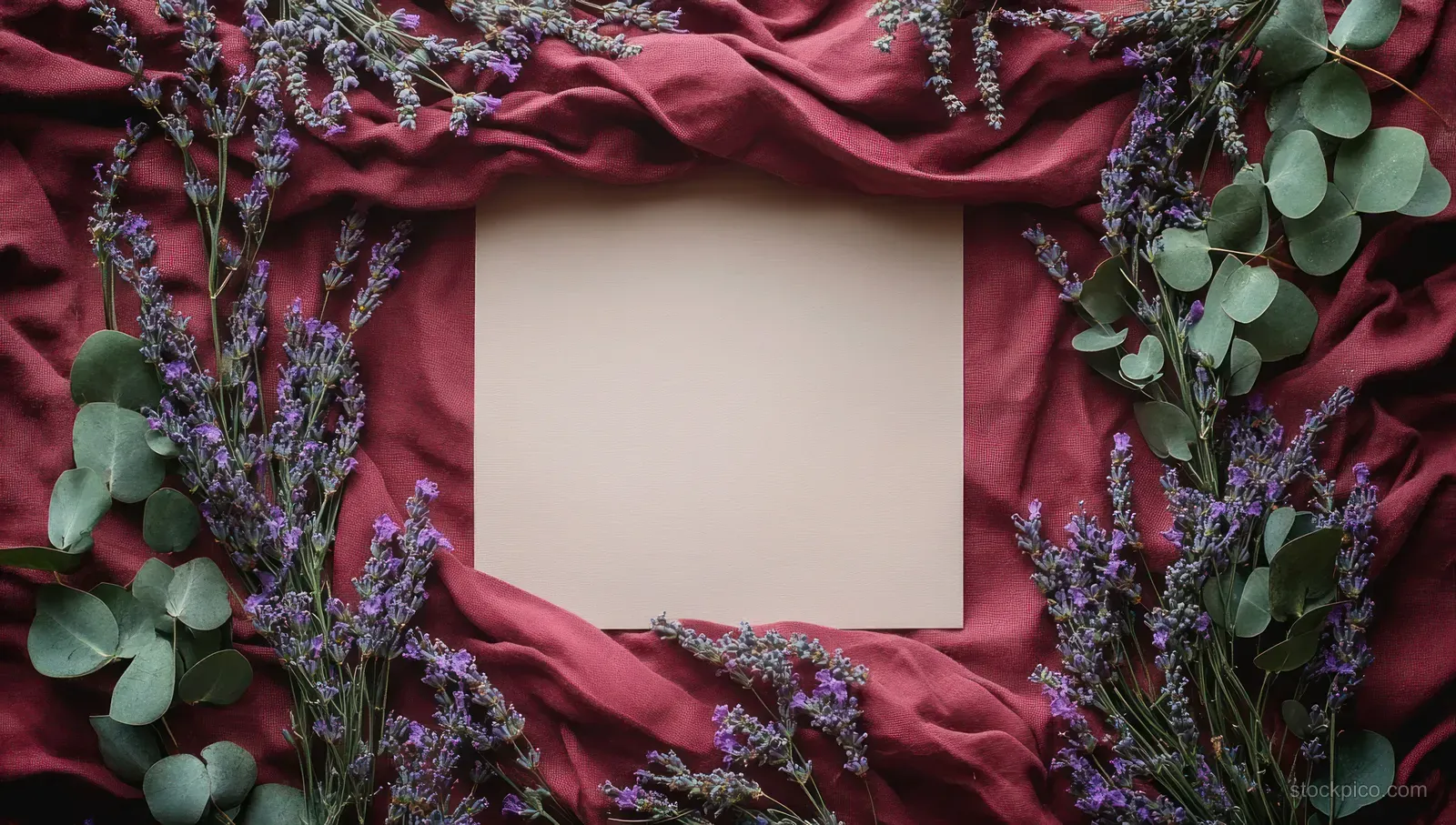Blank beige paper centered on wrinkled burgundy fabric framed by purple lavender flowers and green eucalyptus leaves.