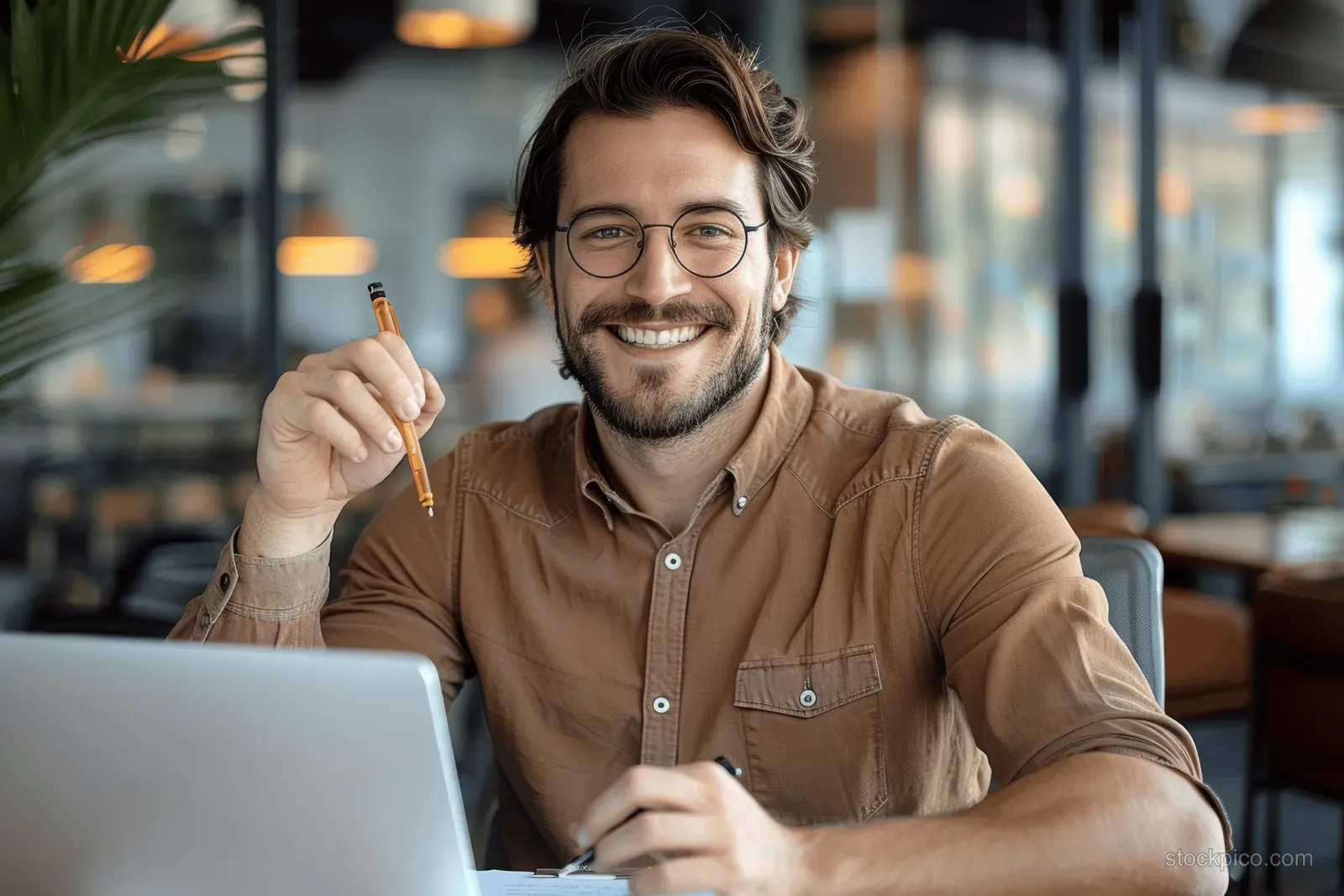 Smiling man in brown shirt sits at an office desk using a laptop with paperwork beside him.