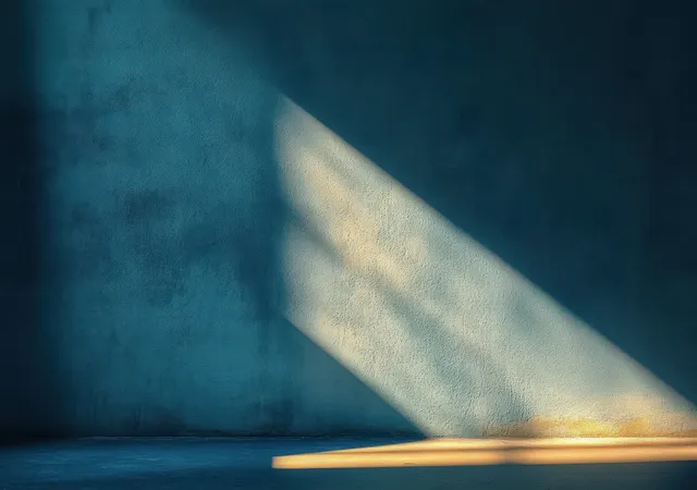 Warm diagonal sunbeam casts sharp shadow streaks across a textured blue wall and empty floor.