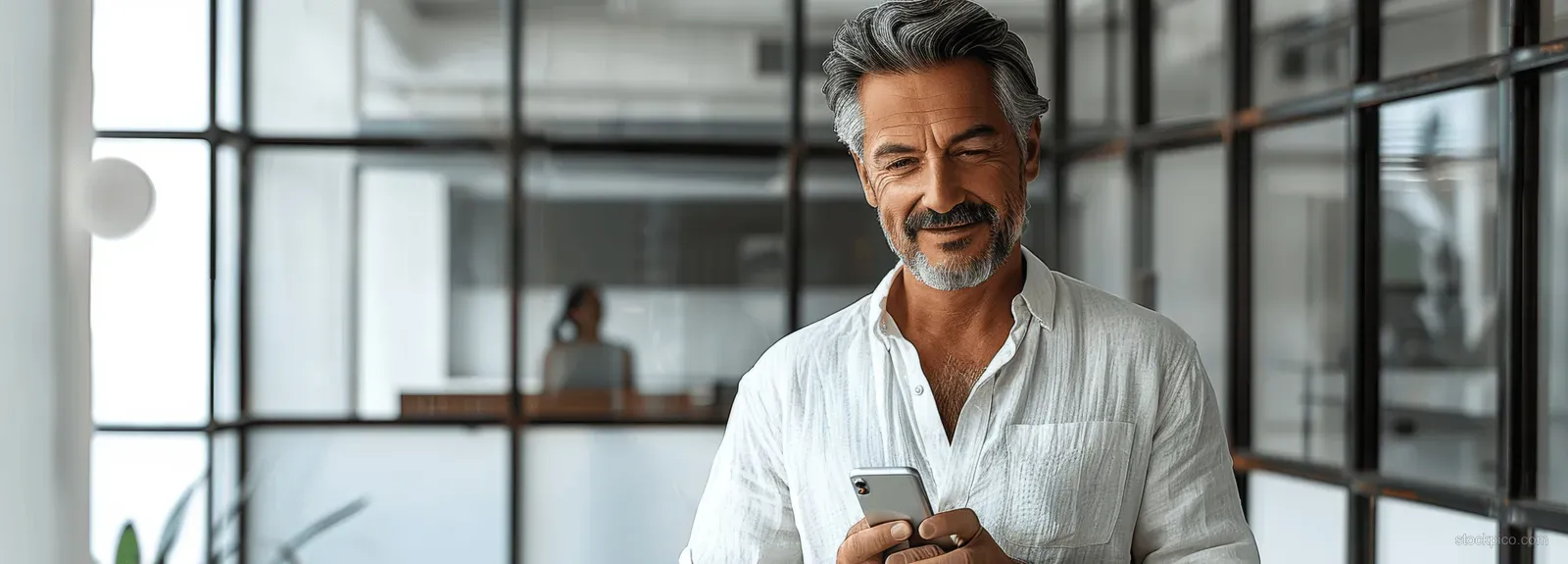 Smiling businessman looking at a smartphone while standing in a bright modern office with glass windows.