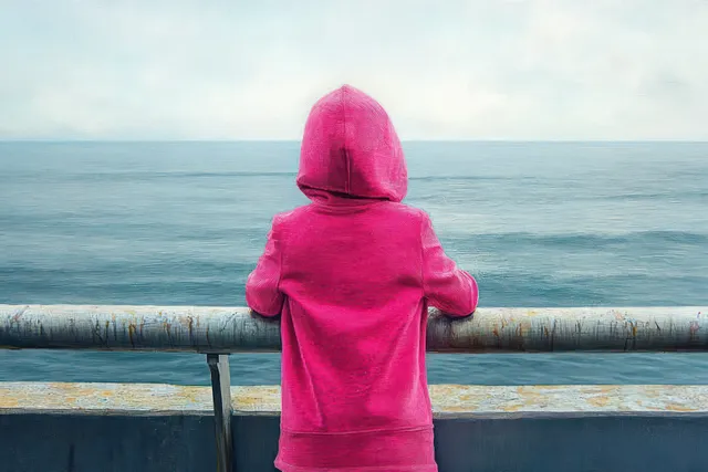 Back view of a person in a bright pink hoodie leaning on a rusty railing overlooking a calm ocean under overcast clouds.