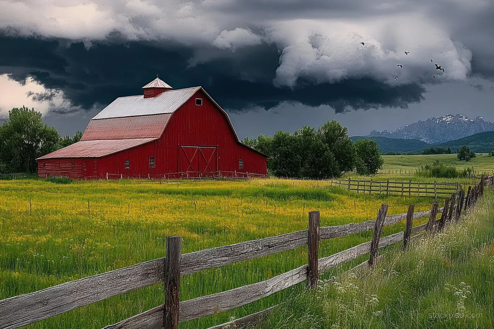 Red barn and wooden fence along a green field under dark storm clouds with distant mountains.