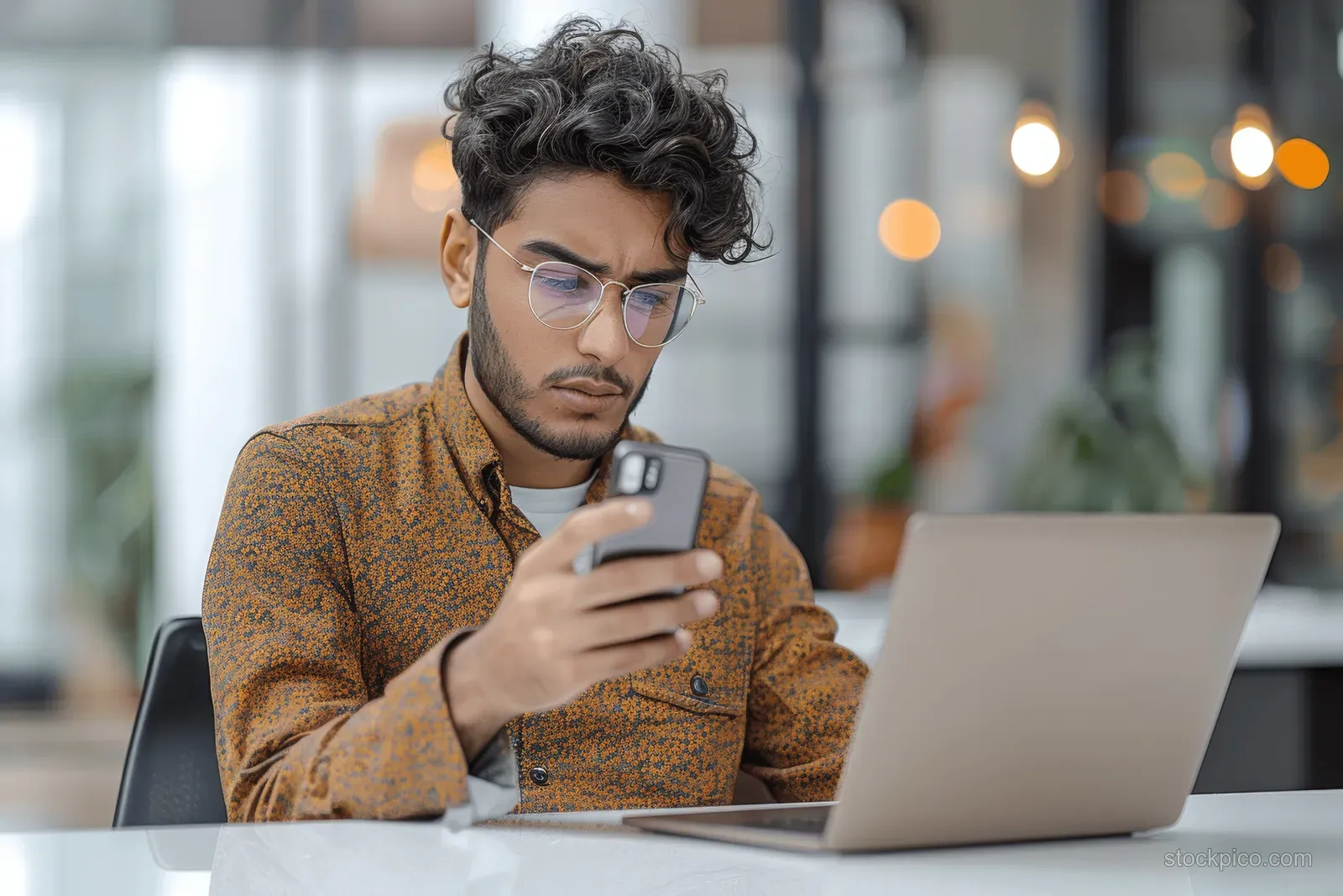 Shocked young man holds a smartphone while working on a laptop at an office desk.