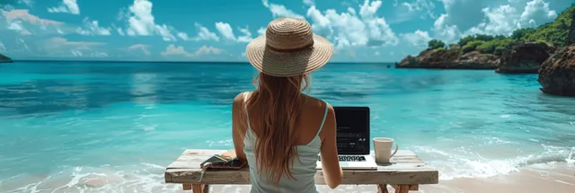 A woman in a straw hat works on a laptop beside a coffee mug at a beach table with the ocean behind her.
