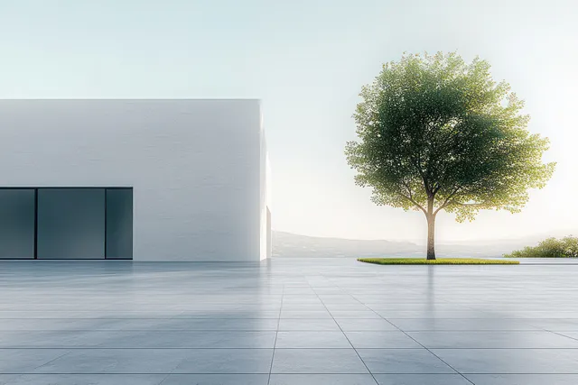 Minimal white building with large glass windows and a single leafy tree on a tiled patio under soft sunlight.