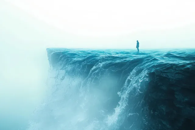 A lone person stands on a wave-textured cliff edge surrounded by mist and fog.