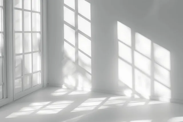 Soft sunlight through window panes casts sharp grid shadows across a minimalist white room interior.