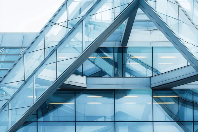 Triangular metal beams frame reflective glass panels on a modern building facade against a bright sky.