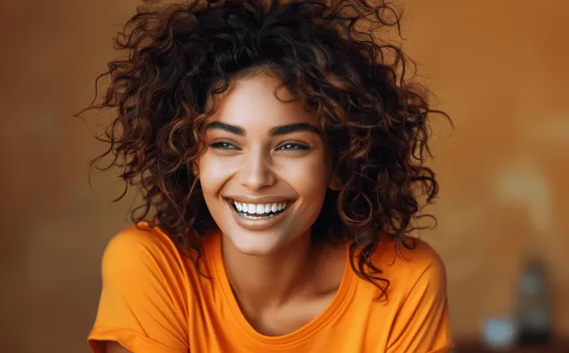 Smiling young woman with curly hair in orange shirt laughing against warm brown background with Smirk To Laughter