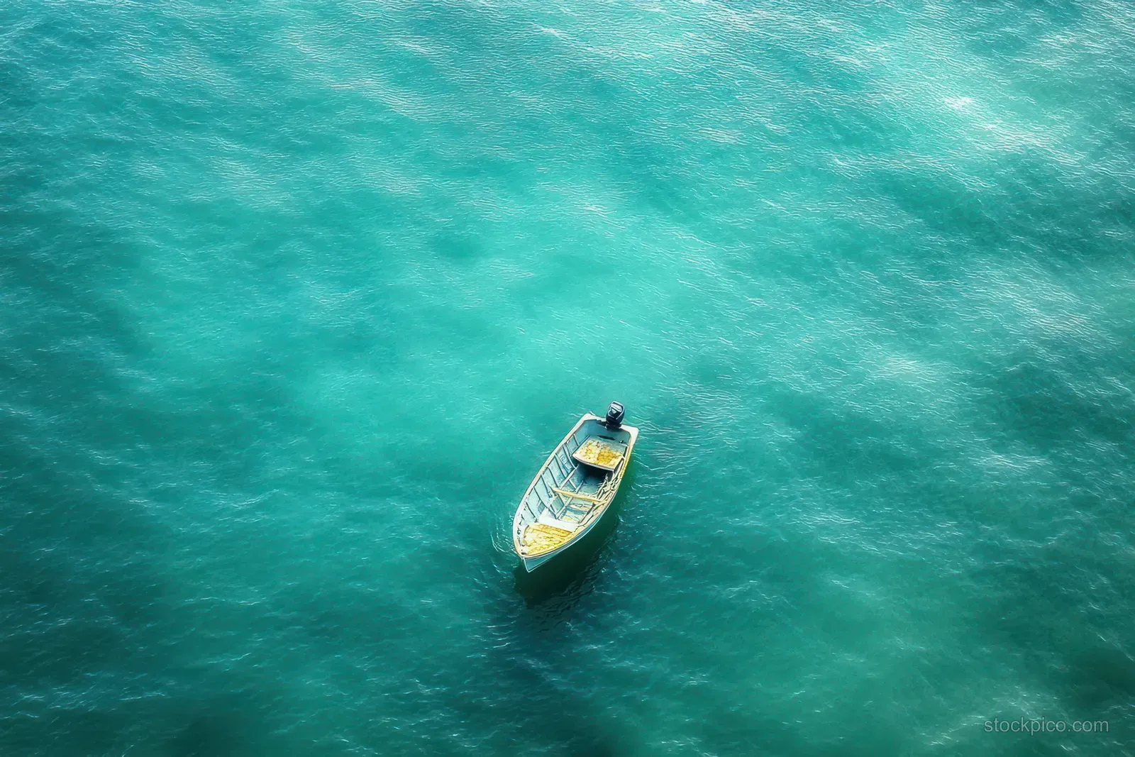 Overhead view of a small motorboat floating alone on calm turquoise water with gentle ripples.