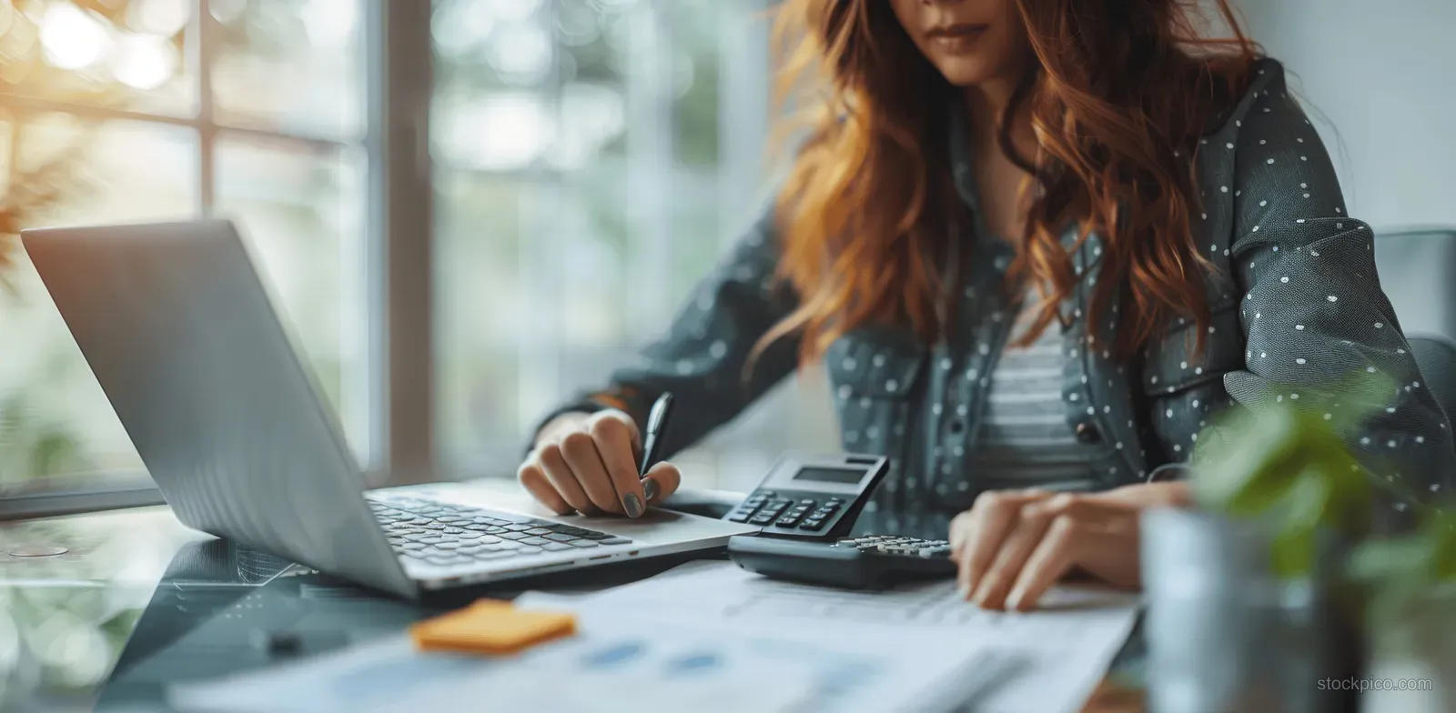 Businesswoman uses a calculator beside a laptop while reviewing financial documents in an office.
