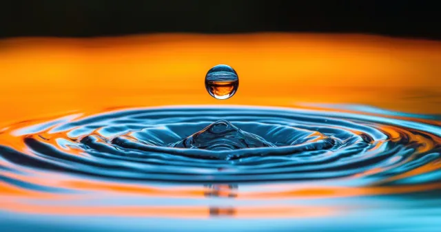 A single droplet suspended above water creates concentric ripples with orange and blue reflected light.