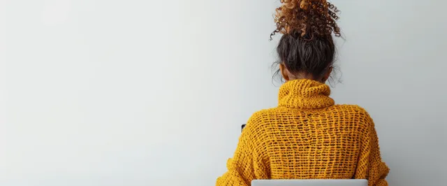 A woman in a mustard sweater types on a laptop at a home desk, shown from behind.