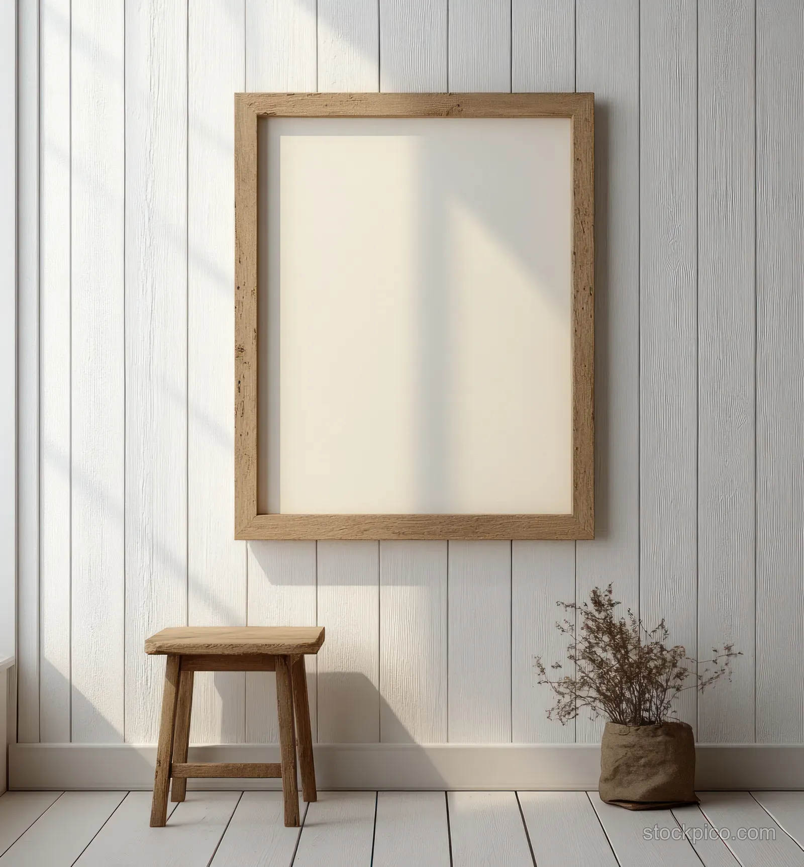 Blank wooden picture frame hangs on a pale shiplap wall above a small stool and potted plant.
