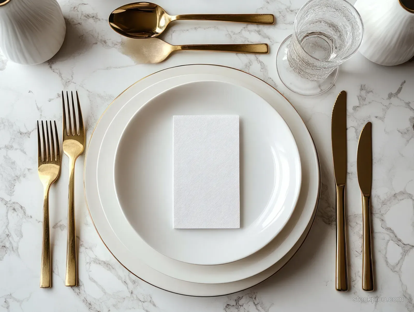 Gold cutlery, white plates, and a clear water glass arranged on a white marble table in a flat lay.