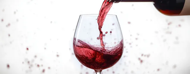 Red wine poured into a clear glass, droplets frozen mid-air against white background