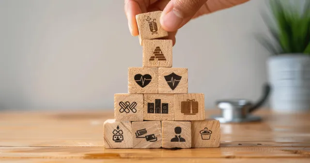 A hand adds a wooden block with insurance and wellness icons to a benefits pyramid on a desk.
