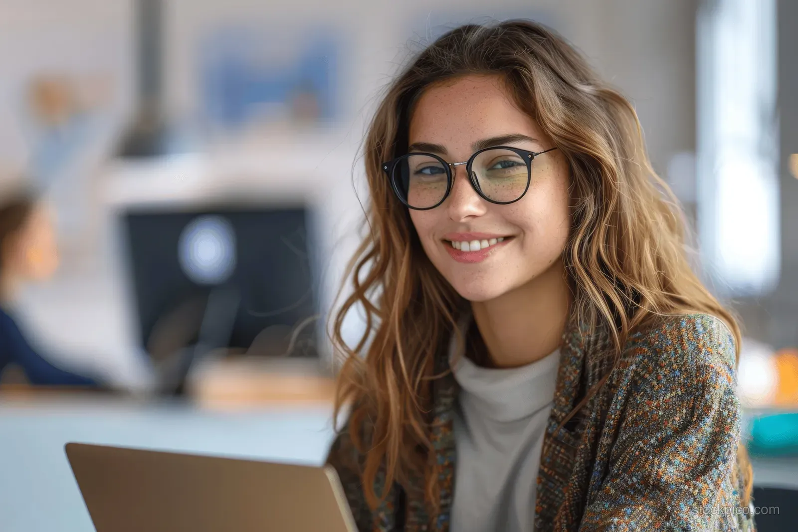 Smiling young woman with glasses using a laptop at an office desk.