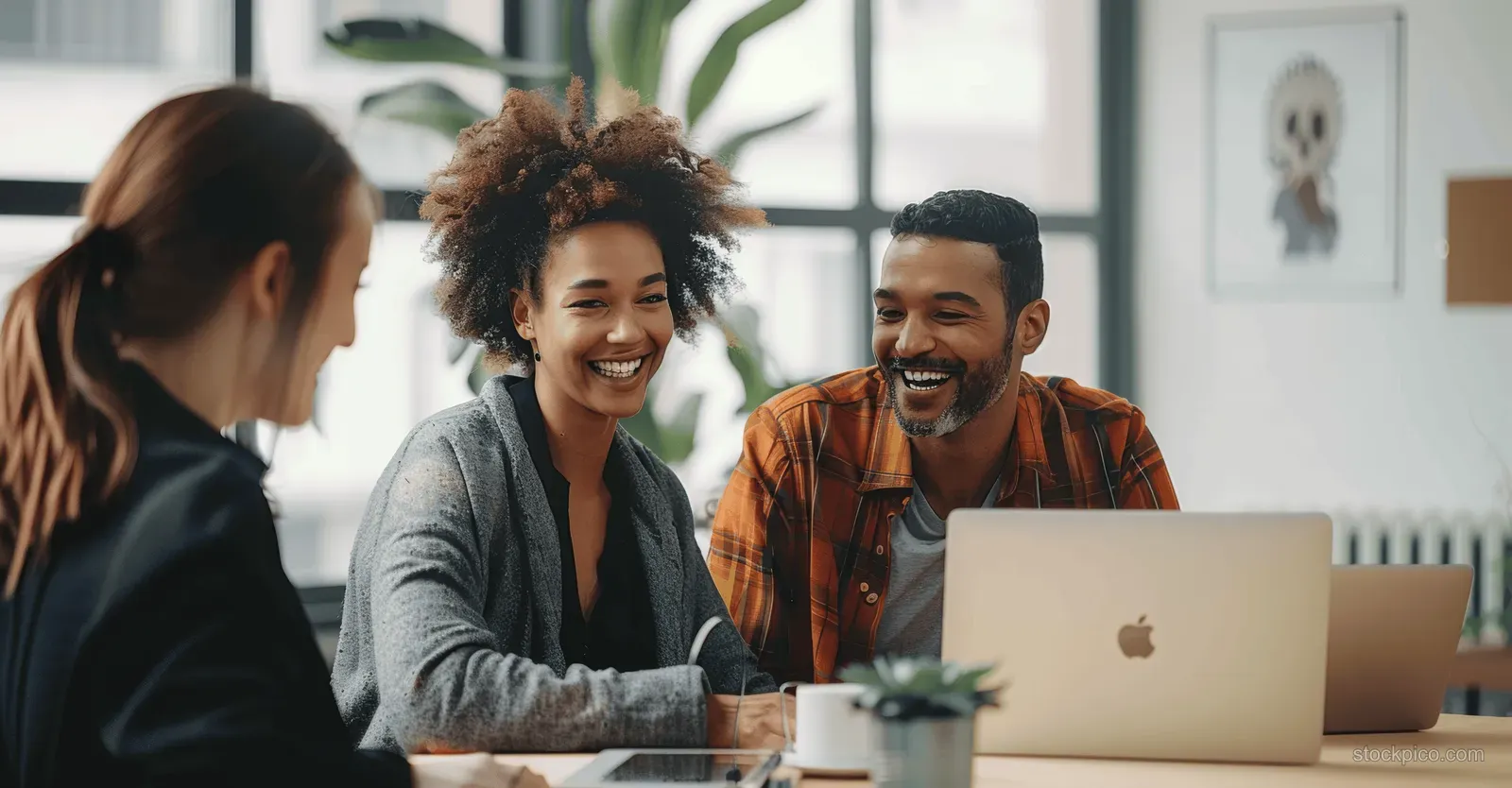 Multicultural coworkers laugh together at a meeting table in a bright modern office while using laptops.