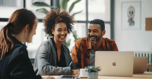 Multicultural coworkers laugh together at a meeting table in a bright modern office while using laptops.