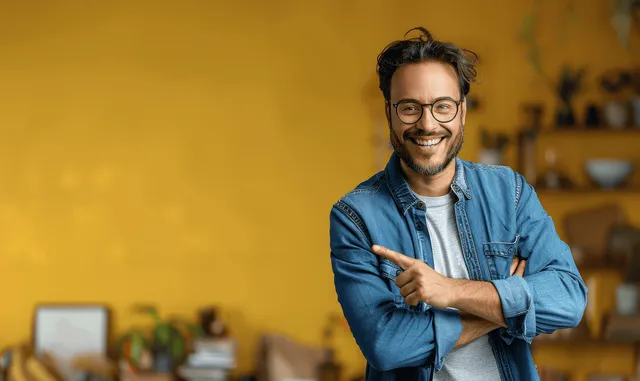 Smiling man in blue denim jacket and glasses points beside empty space on a bright yellow background.