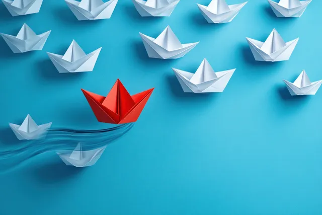 A red paper boat sails away from a group of white boats against a blue background, symbolizing leadership and creative change.